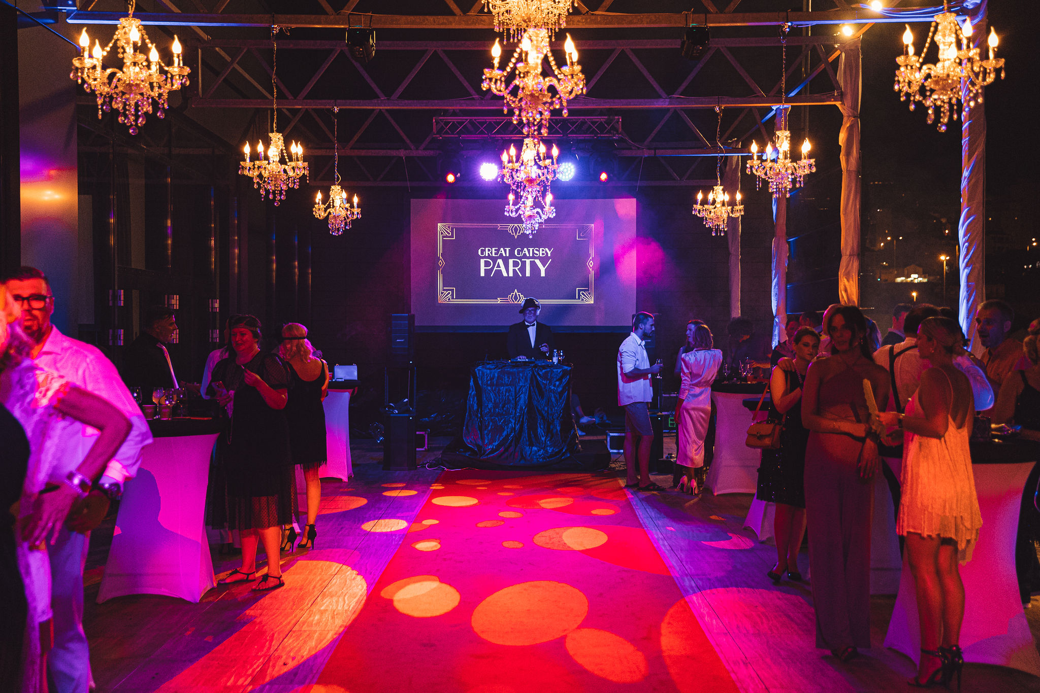 Full venue production at Great Gatsby themed party with LED screen, DJ in tuxedo, crystal chandeliers on truss, gobo patterns on floor, purple lighting, and guests in 1920s costumes