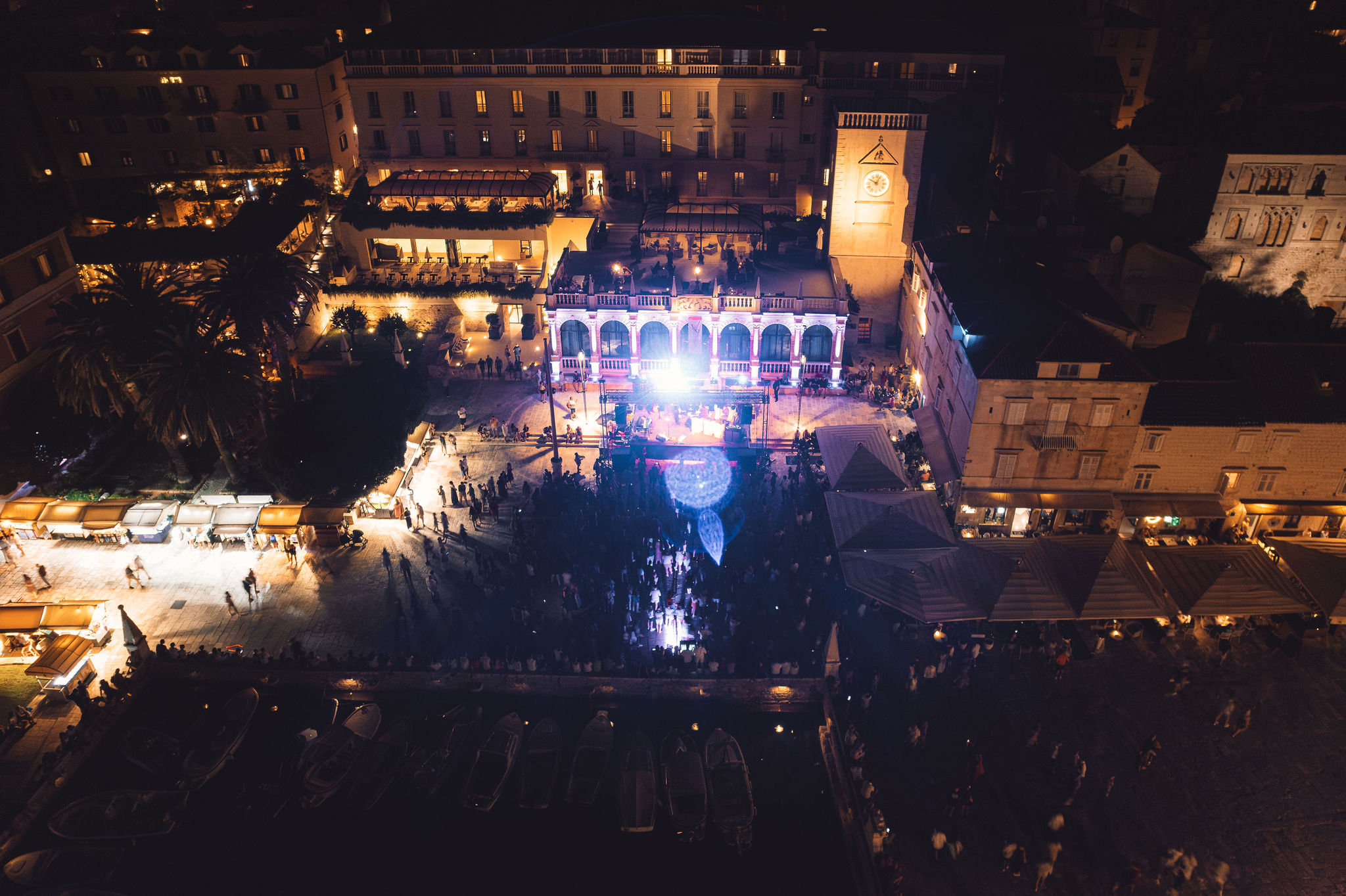 Aerial panorama of Hvar Town square concert at night showing full stage production, crowd, and harbor